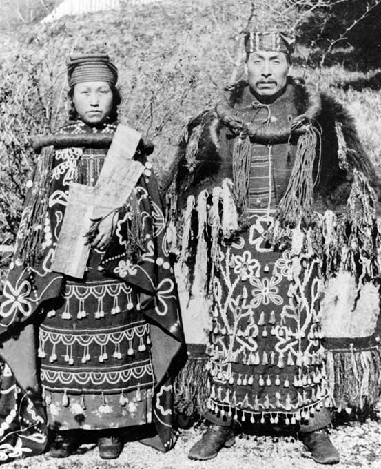 Couple stand side by side in cedar bark regalia, dance aprons, button blankets, she holds a copper in front of her chest.