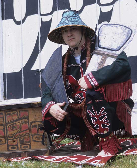 Un homme portant une couverture à boutons et un chapeau d'écorce tressée tient deux Cuivres dans ses mains, accroupi devant une maison cérémonielle.