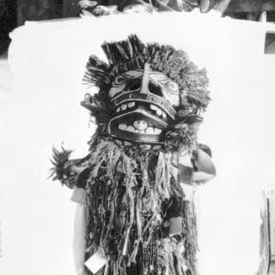 Figure is standing before a white backdrop, body covered with cedar bark fringes, wearing a mask that conceals his face.