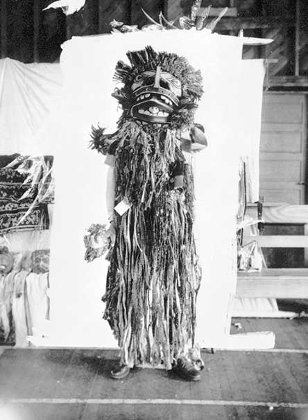 Figure is standing before a white backdrop, body covered with cedar bark fringes, wearing a mask that conceals his face.