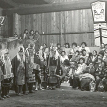 Black and white photograph shows large group of potlatchers wearing ceremonial regalia in a big house.