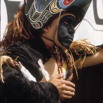 Young male dancer in black face paint wearing regalia comprised of a fur cape and thunderbird frontlet with cedar bark trim.