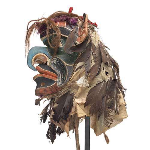 Bakwas mask, beak-like blue hooked nose, perforated brass disk for eyes surrounded by dark green patches, red, white and black facial markings, head covering of feathers mixed with hair, brightly coloured ribbons and cloth, sharp pointed ears.
