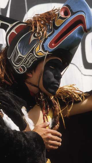 Young male dancer in black face paint wearing regalia comprised of a fur cape and thunderbird frontlet with cedar bark trim.
