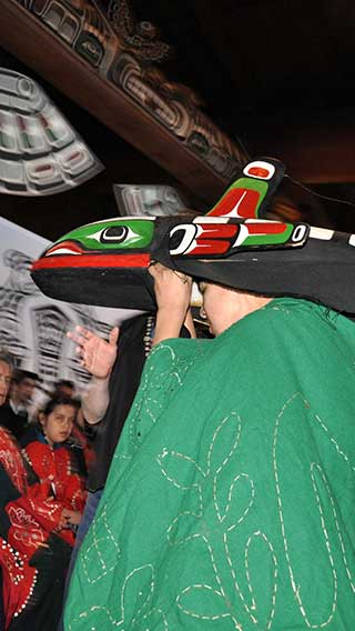 Colour photograph of dancer in whale mask, green robe, potlatch setting in big house, totem pole in background.