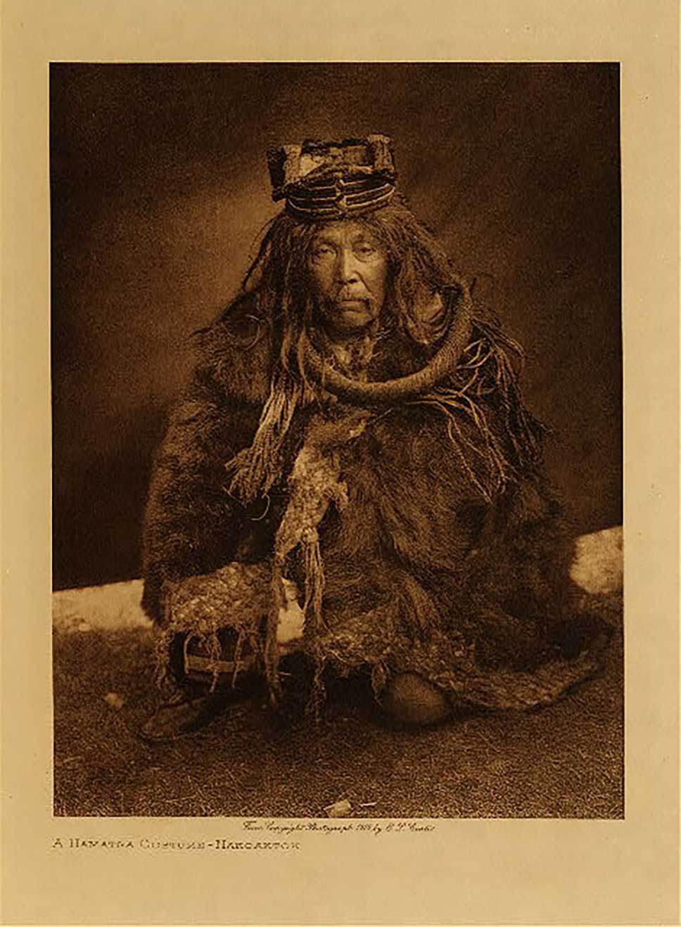Photographie sépia par Edward Curtis un danseur Hamatsa apprivoisé, en tenue d'écorce de cèdre, accroupi, l'air sérieux, en 1910