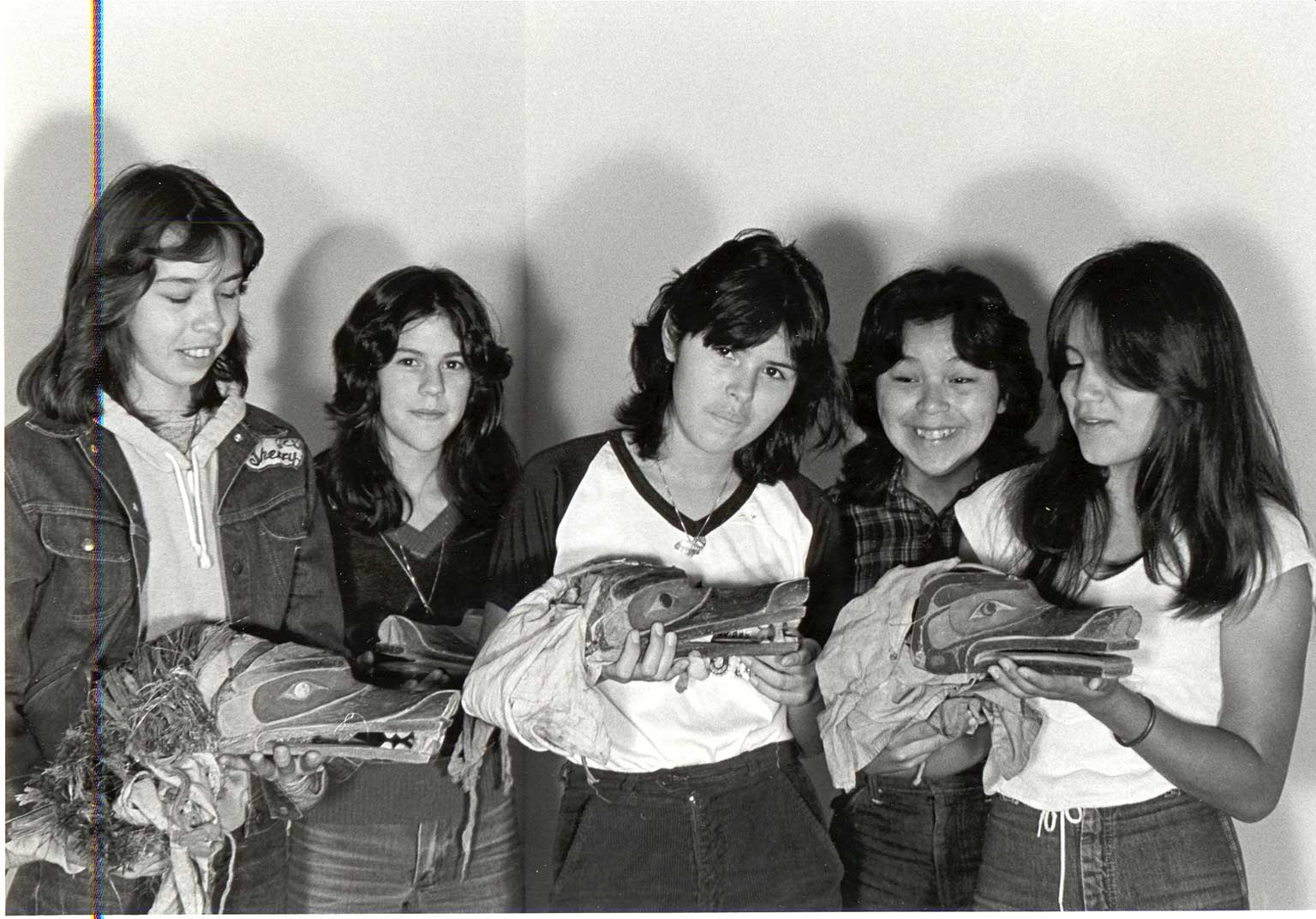 Black and white photograph of five young women in jeans and t-shirts hold 3 wolf headdresses which they observe with pride.