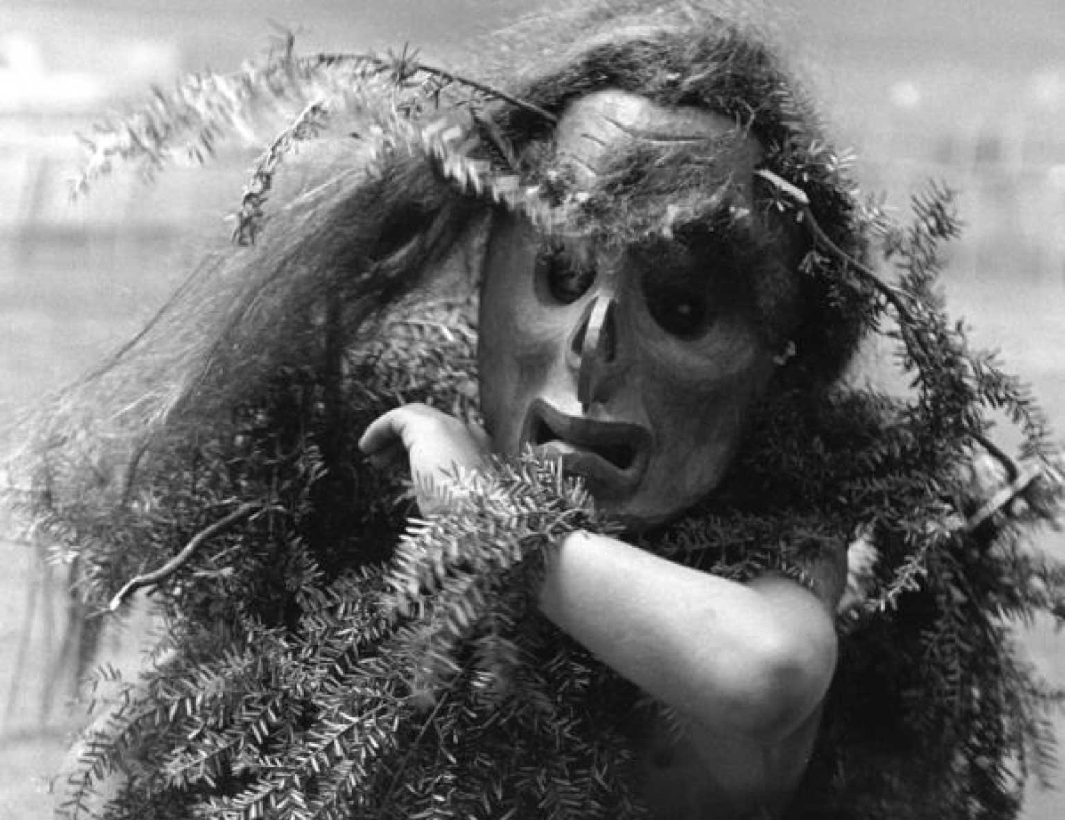 Photographie sépia montrant un danseur Bakwas portant un masque, des branches de cèdre décorent sa tête et ses poignets, un bras relevé cache son visage