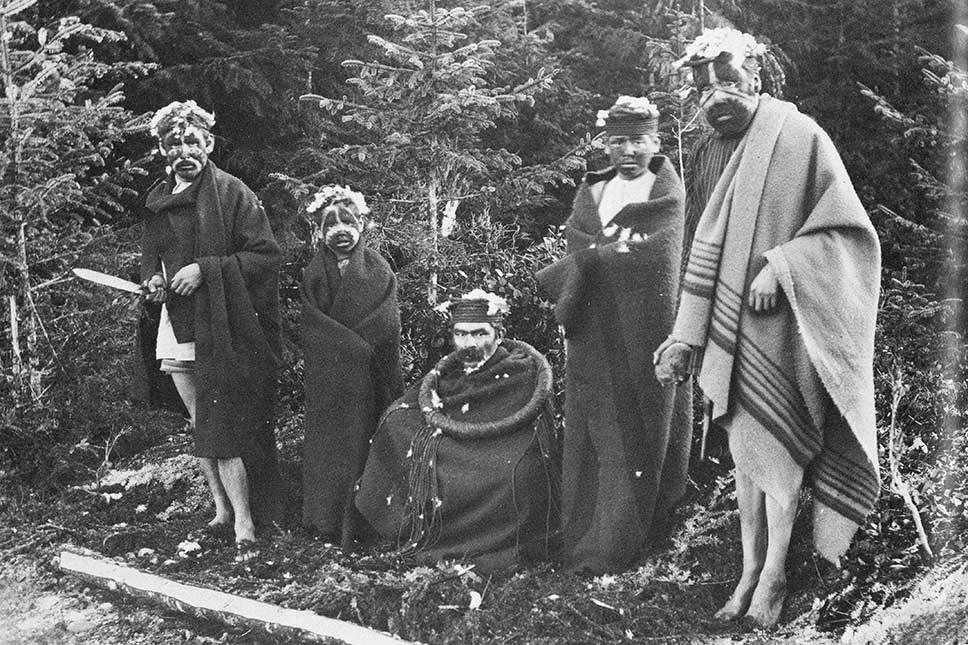 Photographie noir et blanc montrant cinq hommes de tous âges dans la forêt, enveloppés dans des couvertures, les visages peints, de l'écorce de cèdre, des plumes et de la fourrure dans les cheveux