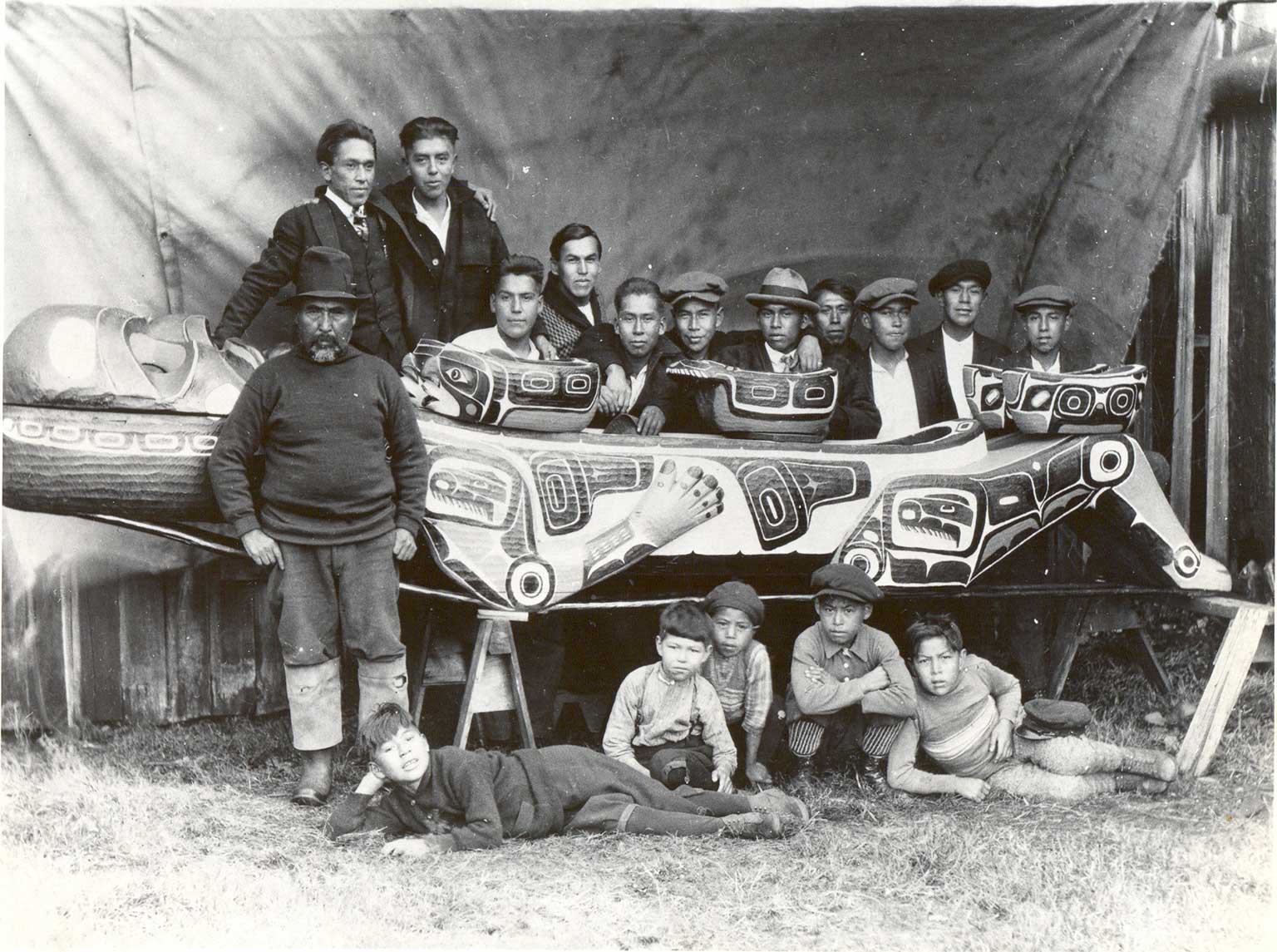 Photographie noir et blanc de garçons assis ou affalés devant un plat cérémoniel géant, derrière le plat se tiennent 12 jeunes hommes vêtus à l'occidentale