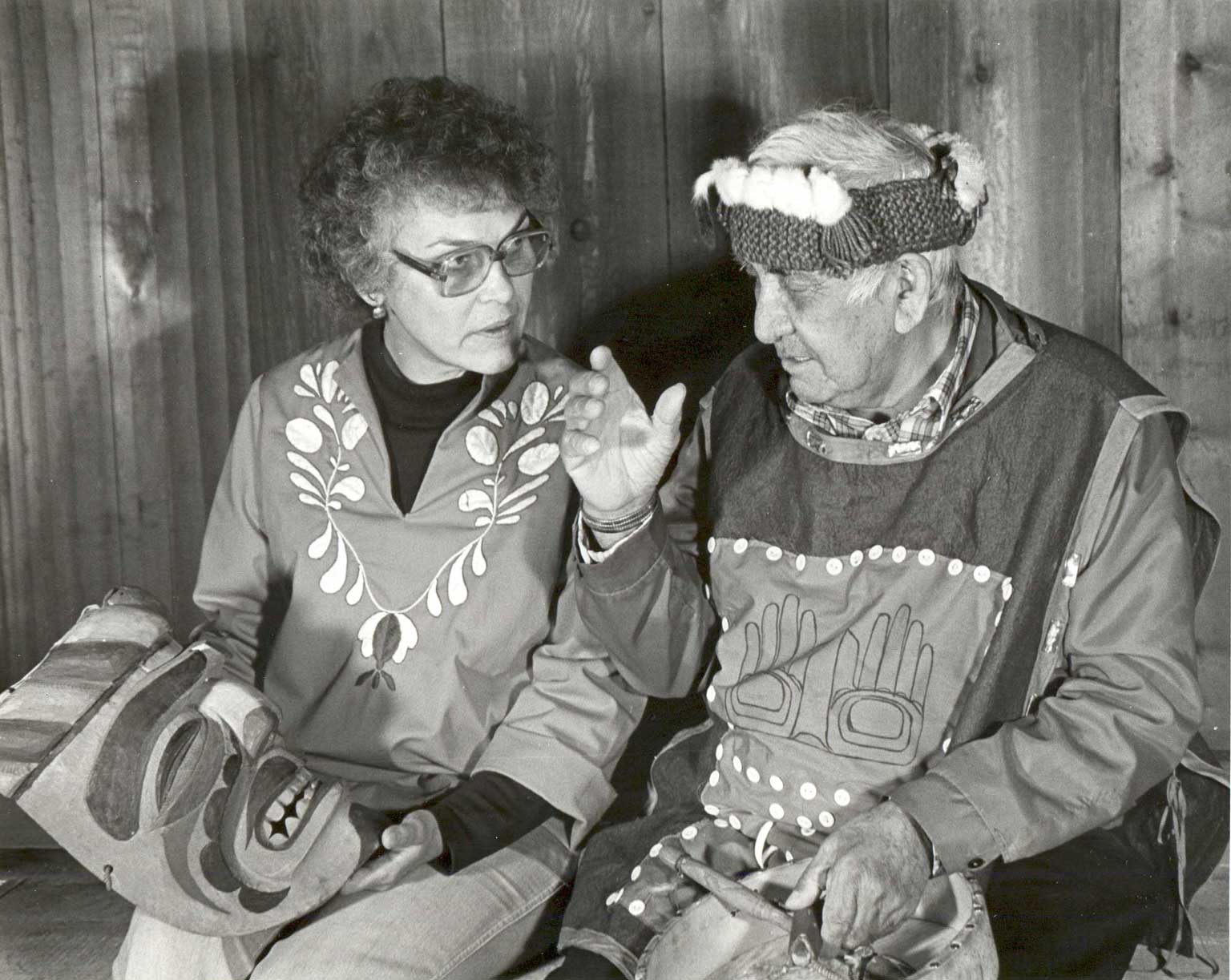 Black and white photograph shows Gloria holding Imas Mask UCC 80.01.017 while Jack - wearing a cedar bark headdress - looks on.