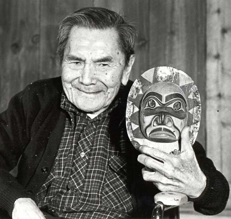 Chief Sam Scow is seated in a wheelchair, holding a frontlet close to his smiling, gentle face.