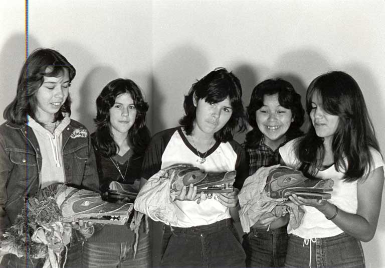 Five young women - casually dressed in jeans and t-shirts - are holding 3 wolf headdresses which they look down upon with pride.