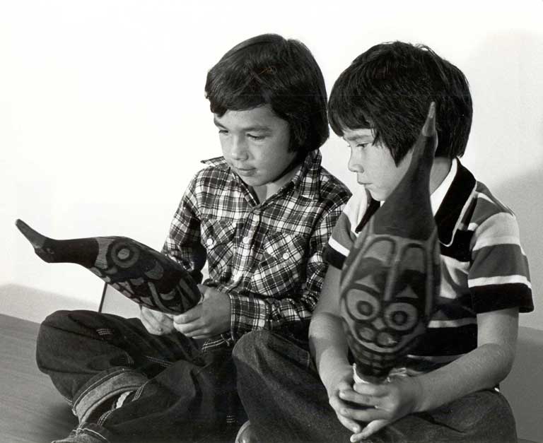 Two young boys are sitting on the floor, each holding a rattle that is shaped like a bird.
