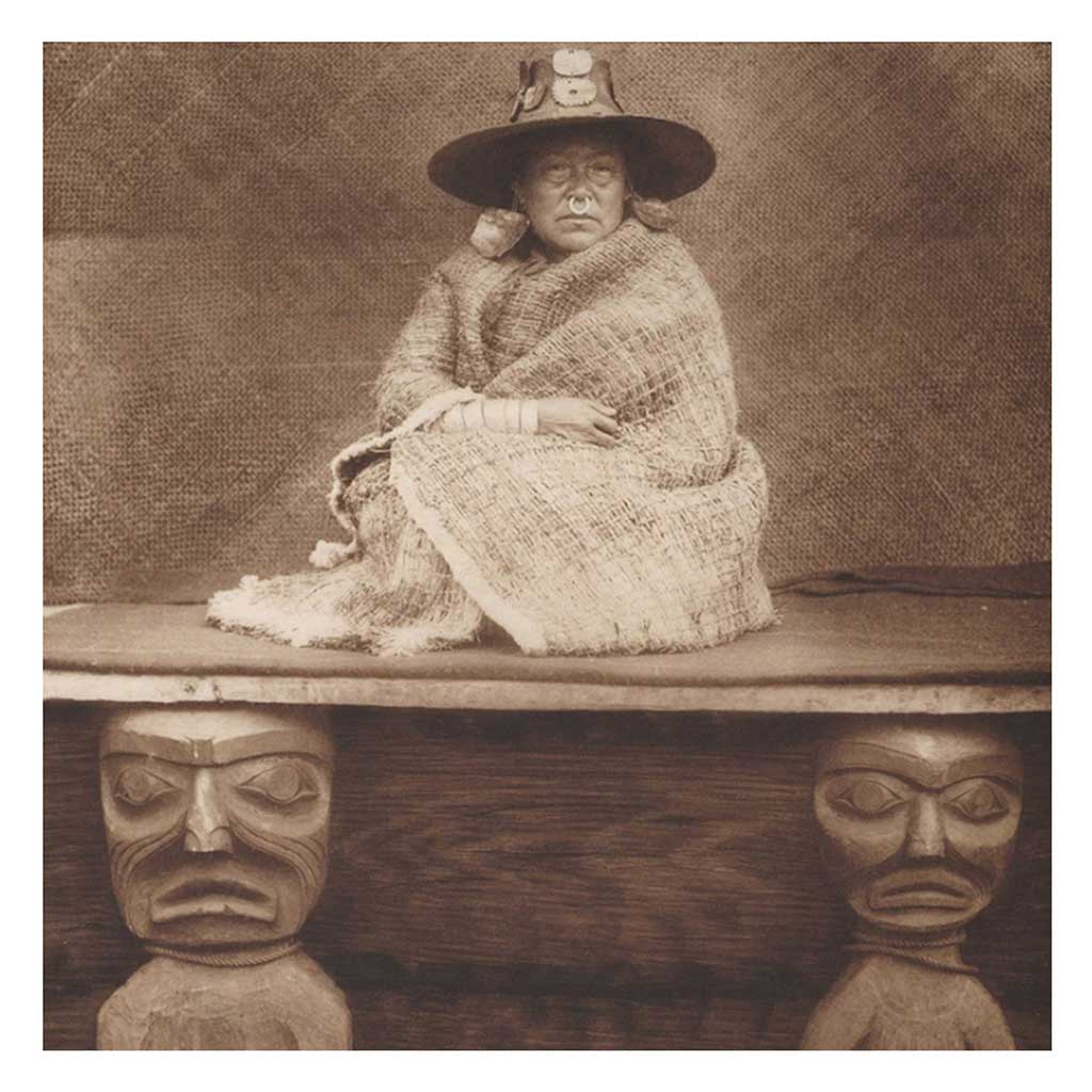 Woman seated on platform above two carved wooden figures. She wears a woven hat, large earrings, nose ring and cedar shawl.