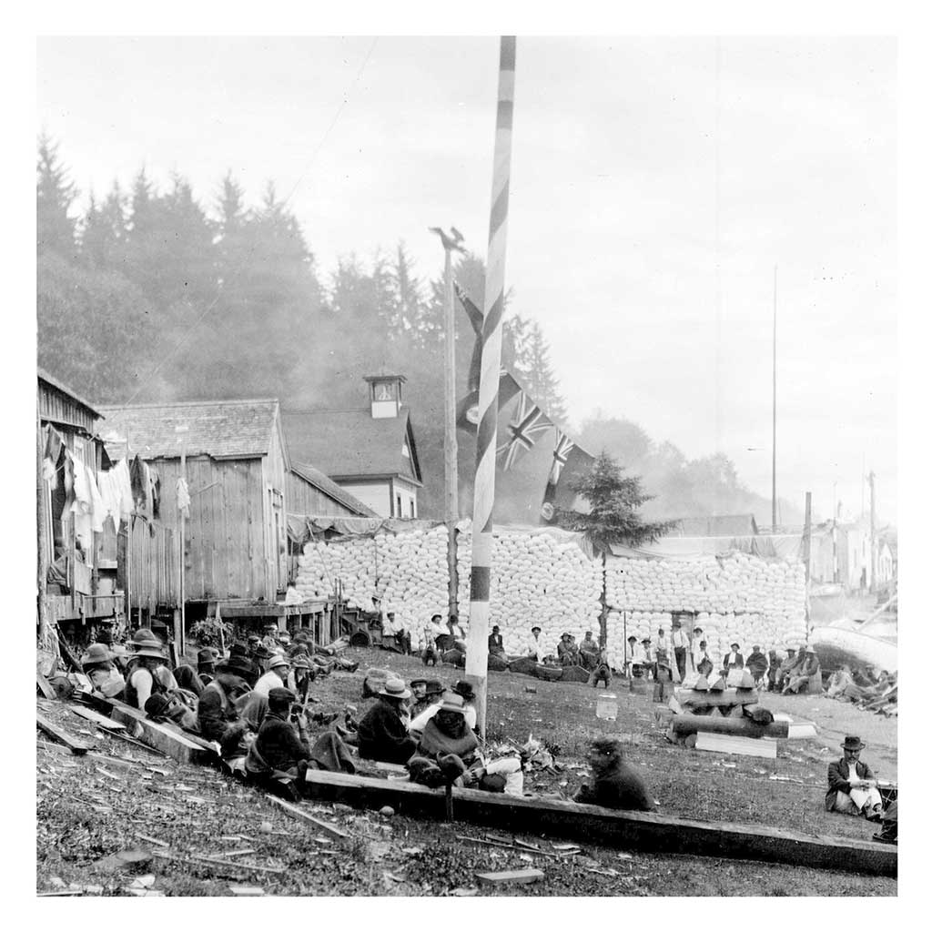 Figures are seated or standing on the beach in front of a large stack of flours sacks with a few small wooden buildings to their left.