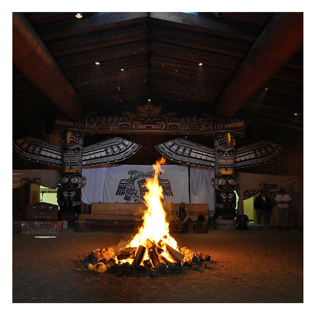Photo couleur prise dans l'obscurité d'une maison cérémonielle, un feu brillant brûlant à l'avant, avec à l'arrière des mâts totémiques et un écran de danse.