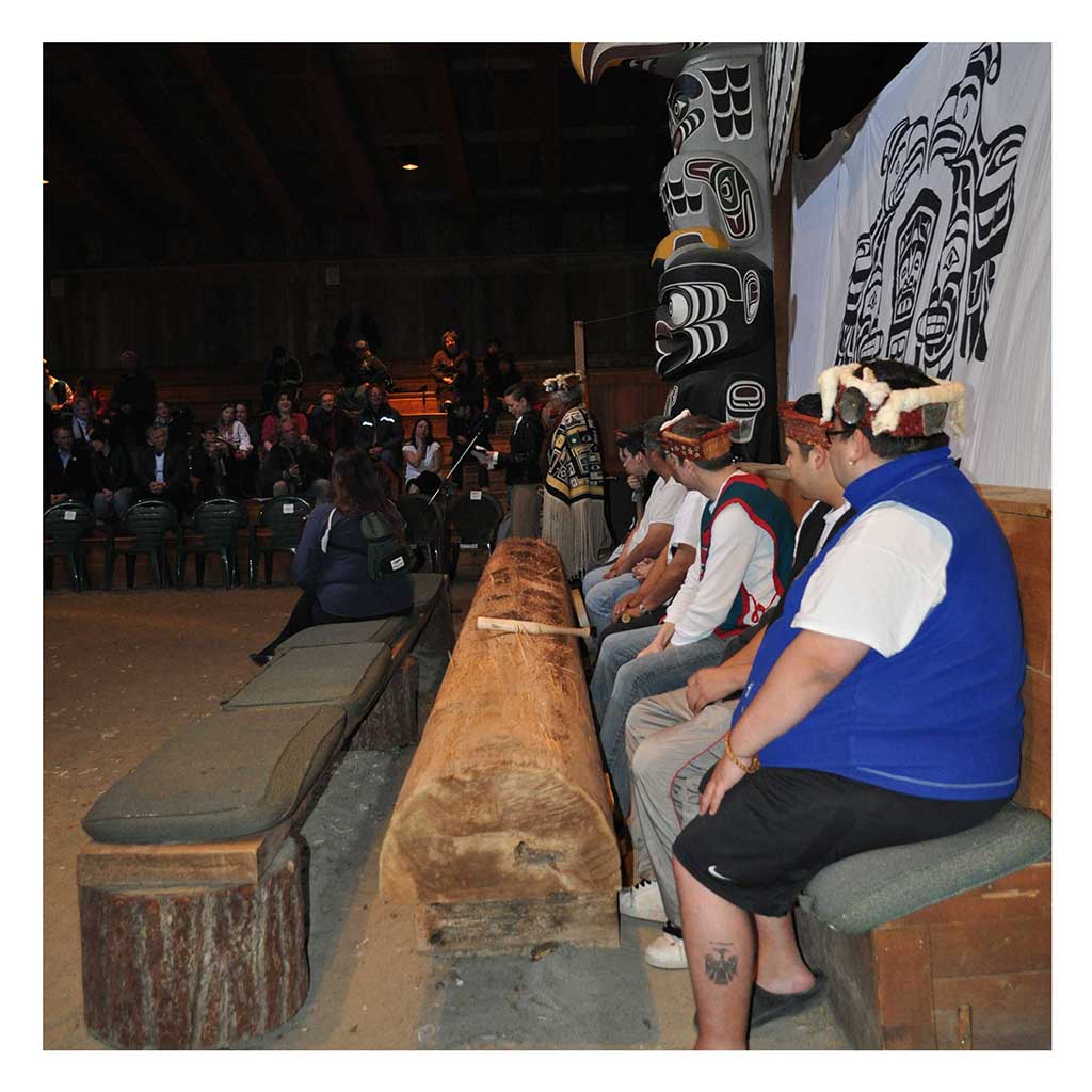 Jeunes hommes assis en rang autour un Tamidzu, un tambour monoxyle, avec derrière eux un mât totémique et un écran de danse.