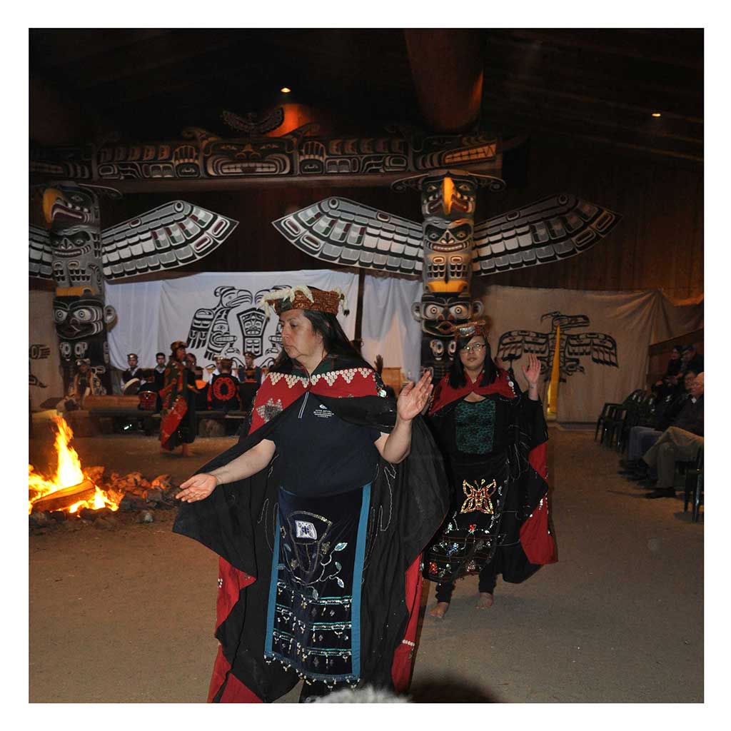 Colour photograph at potlatch in big house of Tłalkwała or ladies dance, two women dressed in dance aprons, button blankets, cedar headdresses.