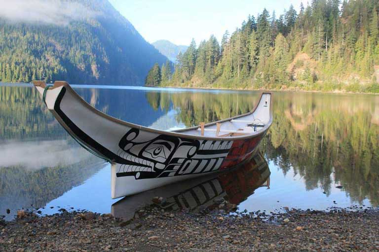 A brightly painted traditional kwakwakw'wakw canoe floating at rest on a beach in lake surrounded by forest.