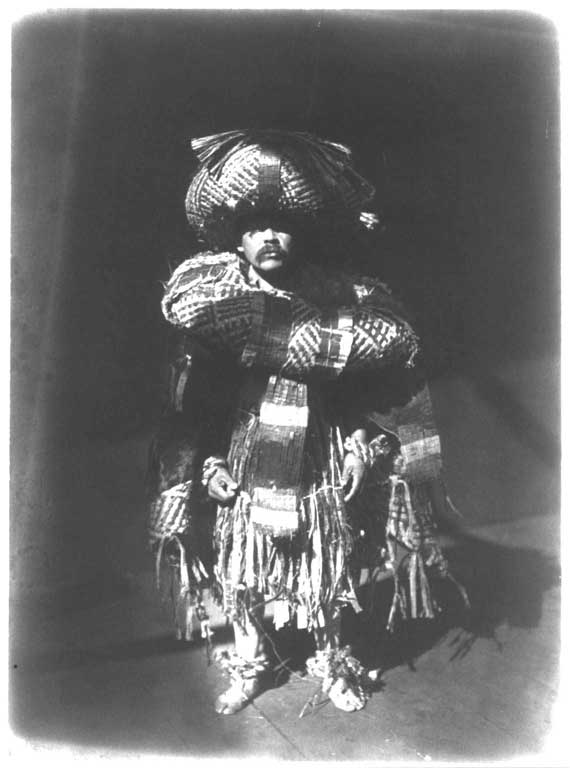 Male dancer appears standing upright, his body covered with cedar bark regalia of hat, neck ring, apron, ankle rings and blankets.