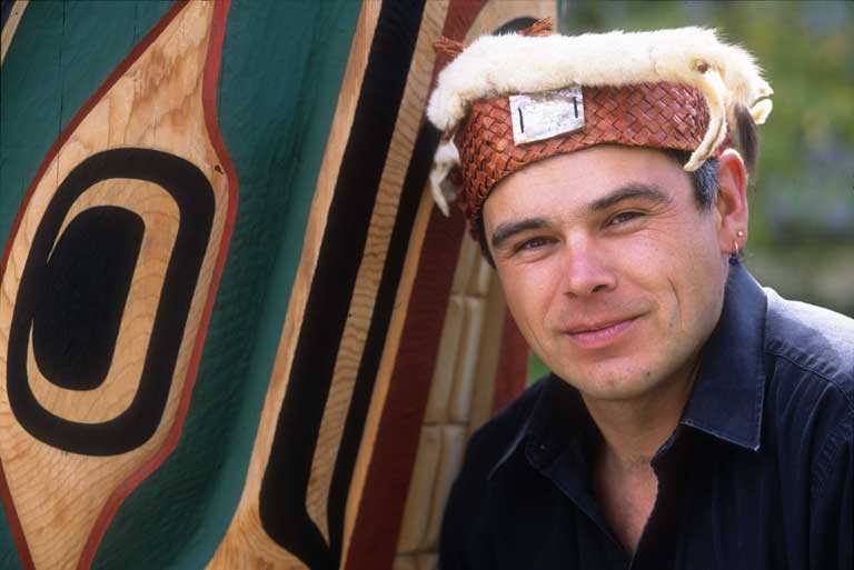 Young adult male wearing a dark blue shirt and traditional cedar frontlet with ermine trim appears before a brightly coloured wooden carving.