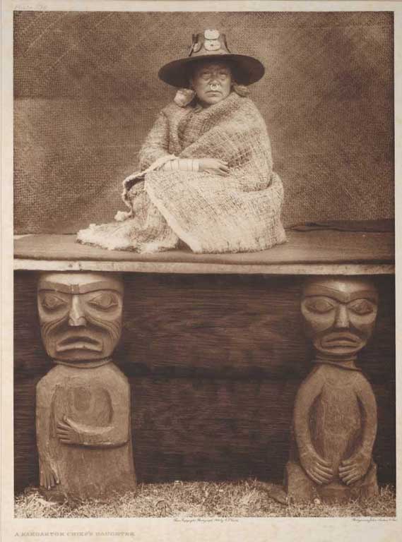 Une femme assise sur une plate-forme aux piliers en bois sculpté porte un chapeau tressé, des boucles d'oreille, un anneau nasal et un châle d'écorce.