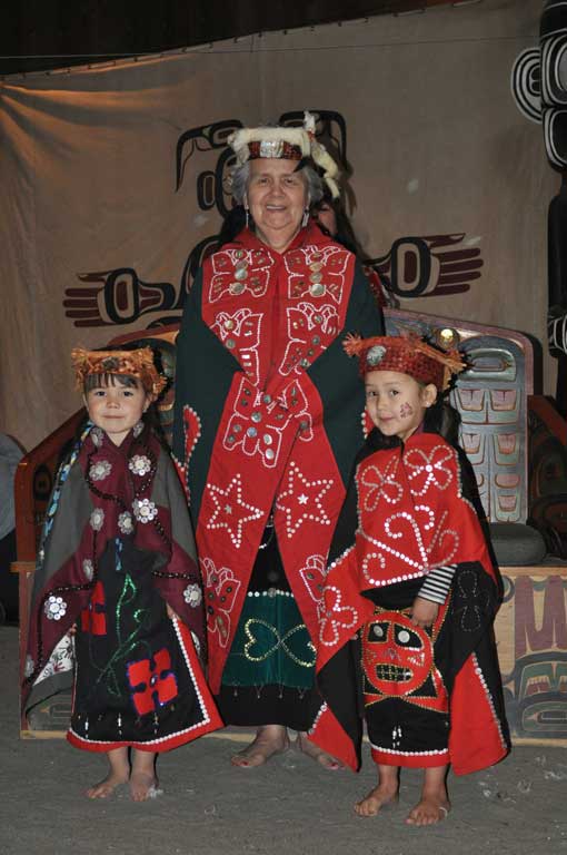 Elder Vera Newman stands in the center of the image with a young girl at each side, all are wearing regalia of button blankets and headdresses.