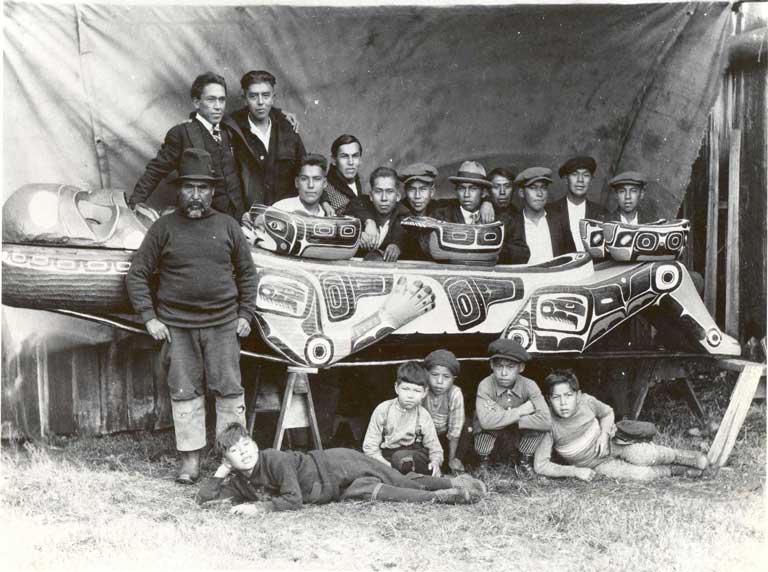 A group of boys lie seated and sprawling in front of a large carved feast dish, behind which stand 12 young men, all wearing western attire.
