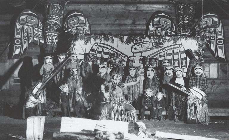 Three men in regalia dance around a fire, behind them a group of masked men stand before a painted screen and totem poles.