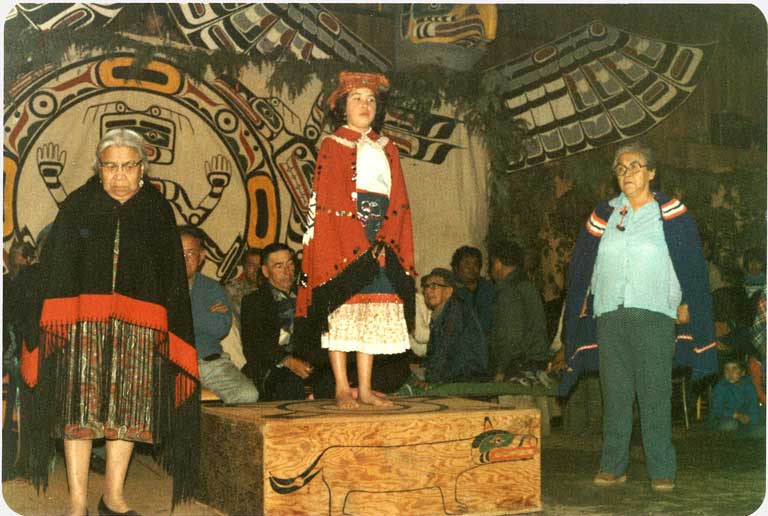 A young girl in a button blanket and cedar headdress stands on a wooden platform before a dance screen with two elders at each side.