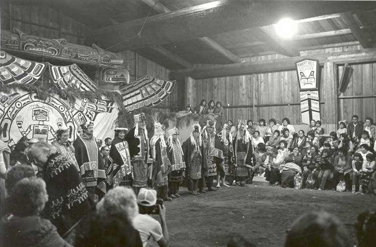 Group of dancers wearing ceremonial regalia stand before brightly painted and carved backdrop along one side of the big house.