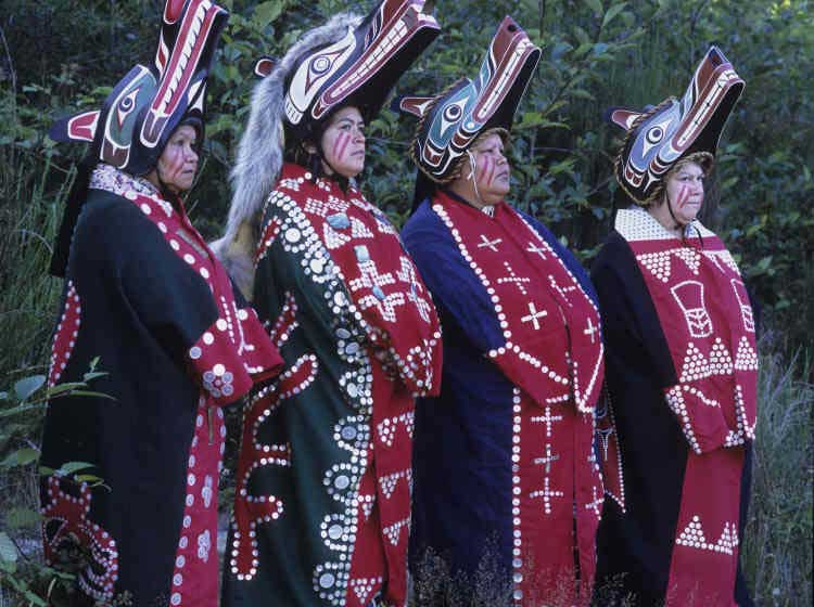 Four women stand in a row facing to the right of the camera, each wears a button blanket and a wolf headdress.