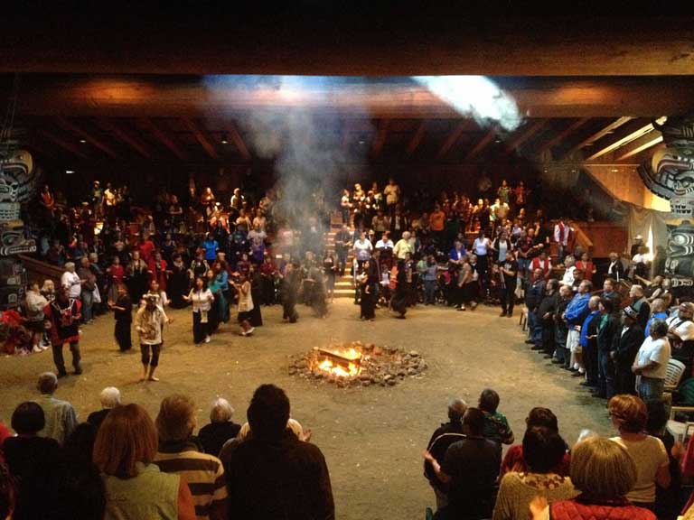De nombreuses personnes se tiennent debout ou dansent autour du feu au centre de la maison cérémonielle.
