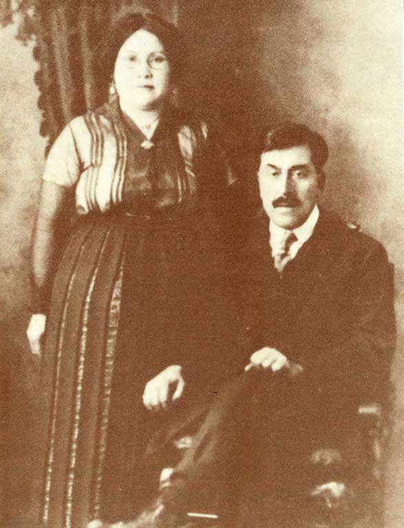 Couple are posed for a studio portrait dressed in western attire, he is seated and she stands at his side.