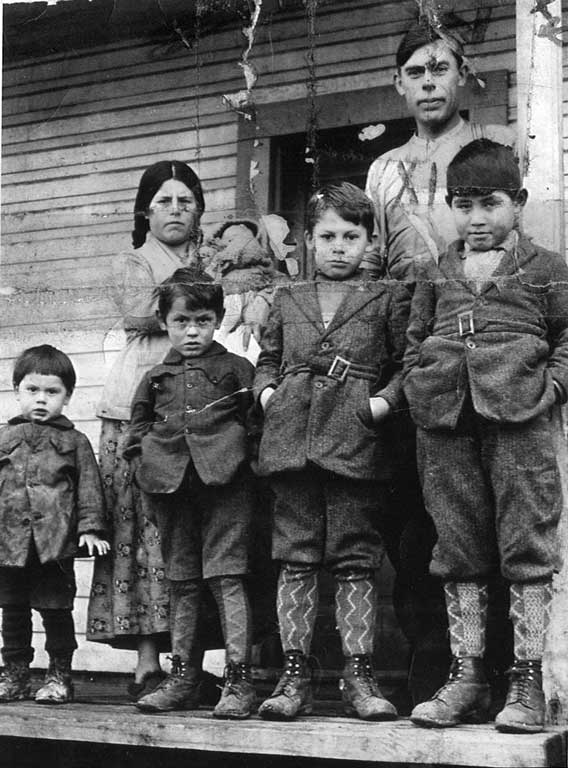 Four young boys in wool coats, knickers, socks leather boots stand before parents, the mother cradles a newborn in her arms.