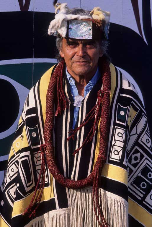 Chief Cranmer appears wearing a Chilkat blanket, cedar neck rings, a frontlet with ermine trim and mother of pearl decoration.