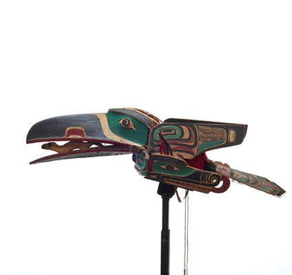 A raven mask, brightly painted with hinged wings, clasping a carved ermine figure in its beak, below which is a sisyutl or serpent figure.