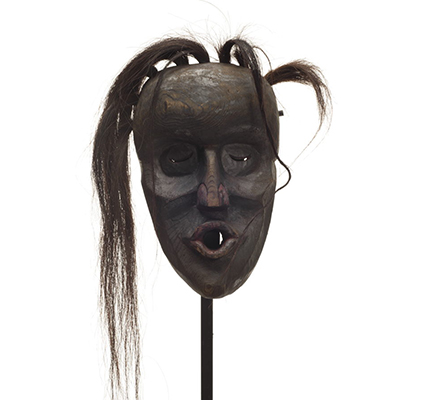 Un masque d'Ogresse Dzunukwa, sculpture profonde, peint en noir, nez et lèvres pincées en rouge foncé, des touffes de cheveux pendant un côté.