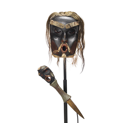 Une dague en bois et un masque Dzunuḱwa de chef profondément sculpté, noir avec des taches rouges, les cheveux clairs.