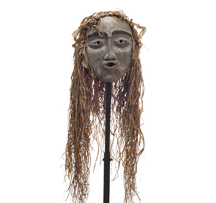 A small forest spirit mask, mostly white face, pursed red lips, prominent black eyebrows, long cedar trim draping from top and back