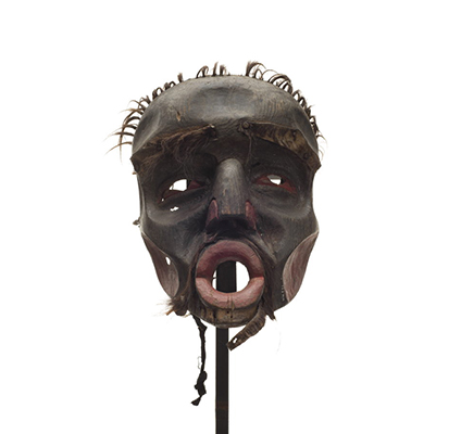 A Dzunuḱwa or wild woman of the woods mask, black stained wood with dark red on nose and cheeks, eyes and nostrils, short tufts of hair on scalp, worn fur patches.