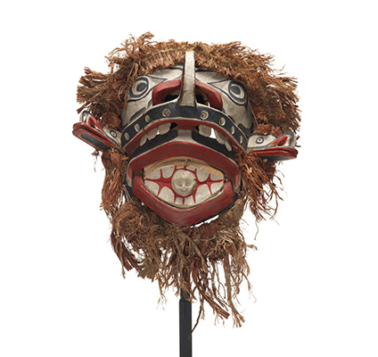 A large and monstrous Baxwbakwalanuksiwe’ mask, surrounded by cedar trim with two mouths, two protruding beak figures and skull figure in mouth.