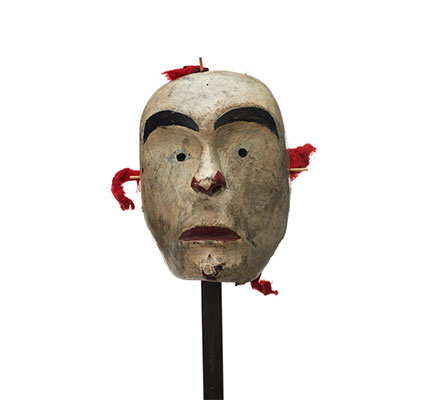 A white-faced mask, small circular eyeholes, red paint on lips and surrounding nostrils, bright red fabric rigging.