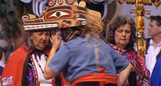 A dancer wearing a wooden hat-like mask appears with her back to the camera, two other women in red capes face the camera.