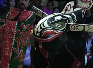A red and green button blanket appears to the left of a brightly painted whale mask with pectoral fins open.