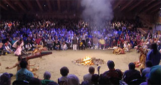 Smoke rises above a burning fire pit in the big house. Around the fire a group of dancers and behind them the audience.