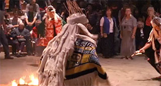 Three dancers in ceremonial regalia move around a fire at a potlatch in the big house.