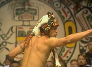 Shirtless man wearing a headdress-style mask stands in profile before a dance curtain and points to the right.