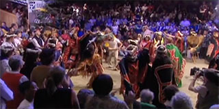 A group of dancers move in circles around the dirt floor at the center of the big house, they are surrounded by audience.